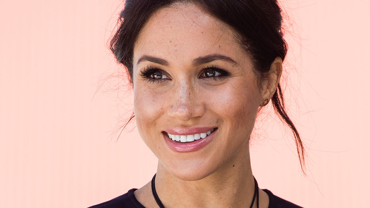 A close-up of Meghan Markle smiling wearing a black top and a black necklace