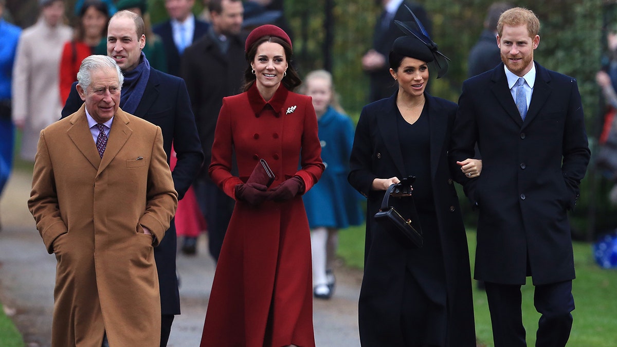 King Charles in a Chestnut colored jacket walks in front of Prince William and next to Kate Middleton in a red jacket, next to Meghan Markle in a black jacket, who holds onto the arm of Prince Harry in a black suit