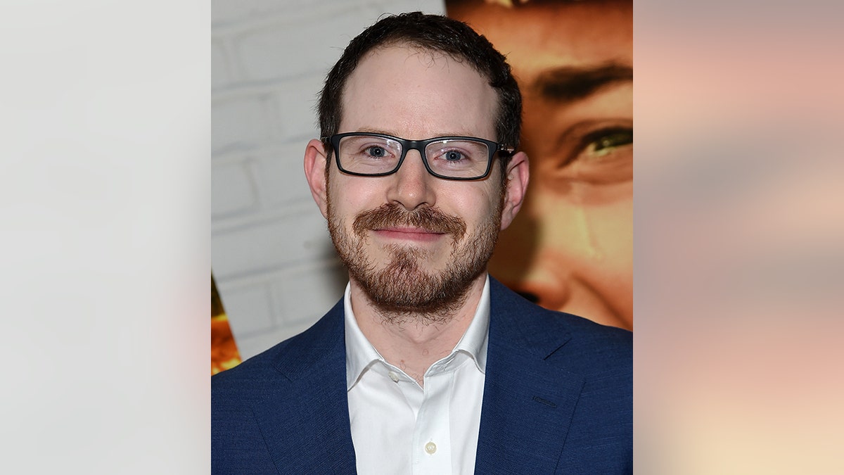 Ari Aster in a navy suit soft smiles on the red carpet wearing dark framed rectangular glasses