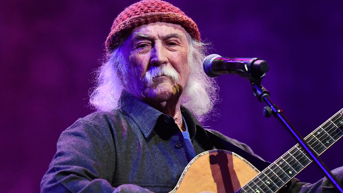 David Crosby on stage holding a guitar, wearing a dark jean shirt and red knitted cap