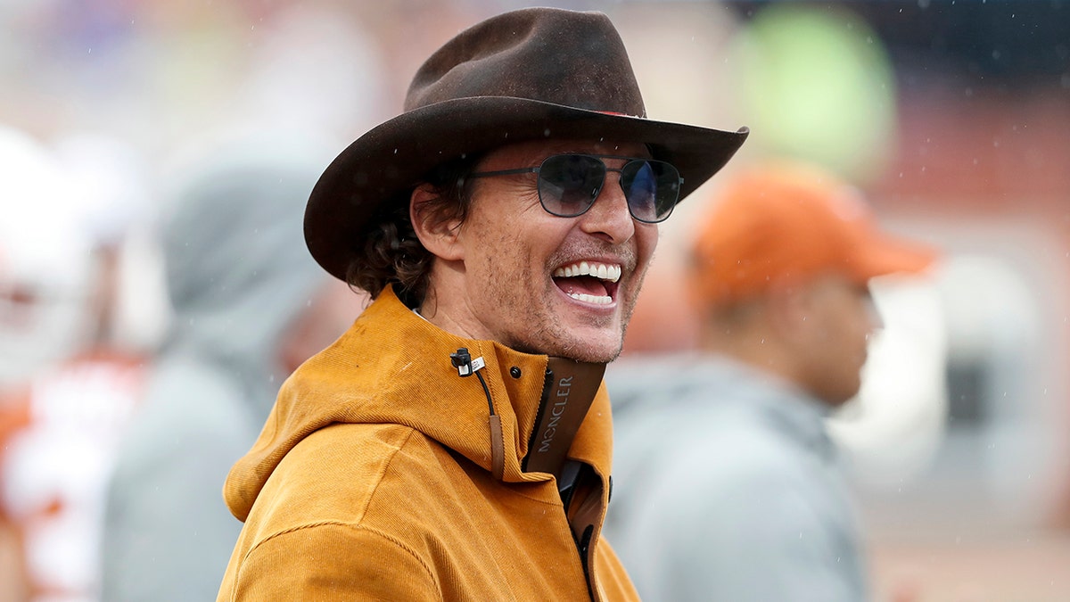 Matthew McConaughey in an orange jacket laughs with a brown cowboy hat on and sunglasses