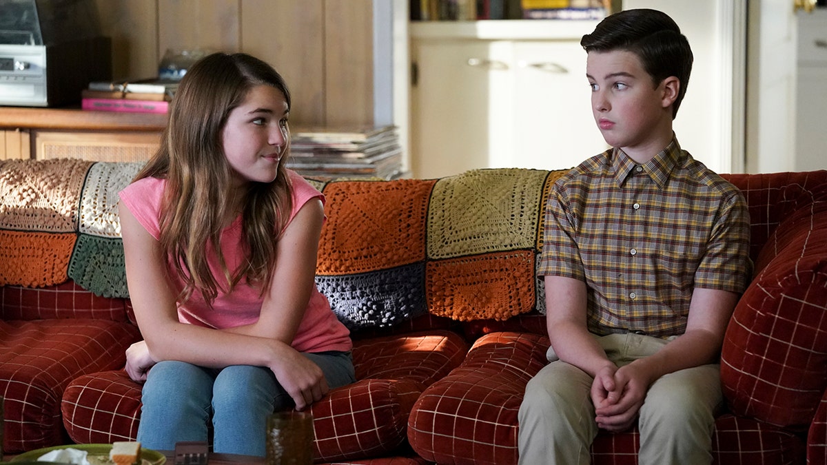 Missy (Raegan Revord) in a pink shirt stares at twin brother Sheldon (Iain Armitage) in a plaid short-sleeve shirt while sitting on the couch during an episode of "Young Sheldon"