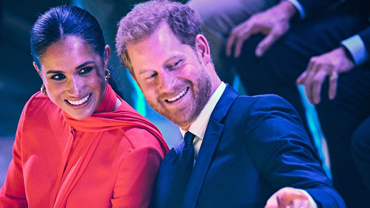 Meghan Markle smiles in a red gown while Prince Harry Leans back towards her and also smiles in a blue suit