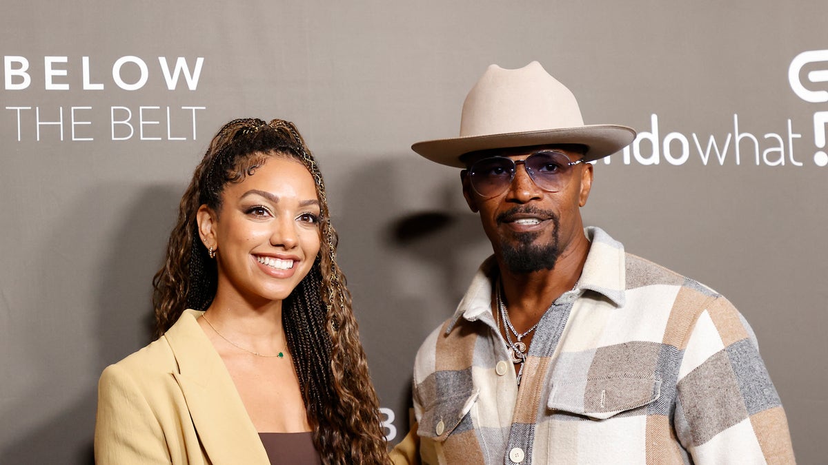 Corinne Foxx in a tan jacket and brown top smiles on the red carpet next to father Jamie Foxx in a patch multi-color shirt with a light tan wide-brimmed hat