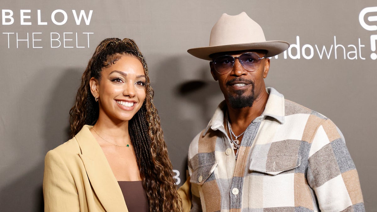 Corinne Foxx in a tan jacket and brown top smiles on the red carpet next to father Jamie Foxx in a patch multi-color shirt with a light tan wide-brimmed hat