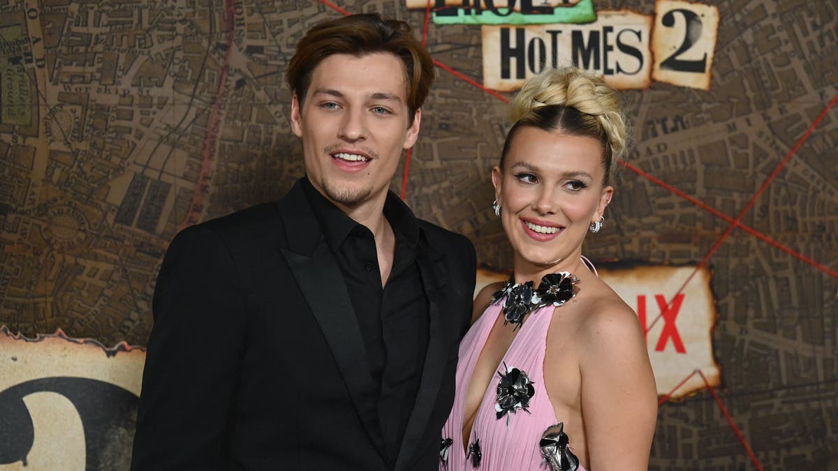 Jake Bongiovi in a black suit poses next to Millie Bobby Brown in a pink dress.