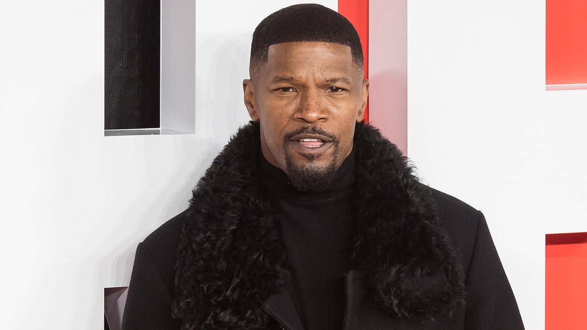 Jamie Foxx in a black turtleneck and coat with a furry trim looks snazzy posing for a photo on the red carpet