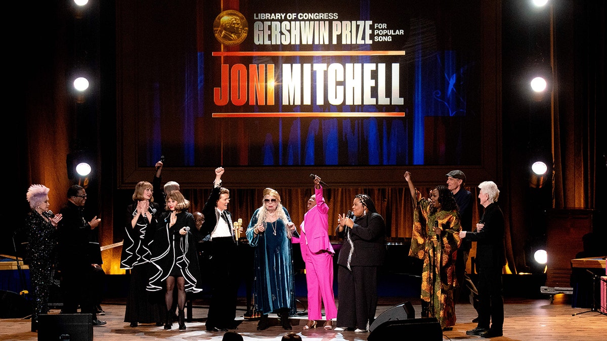 Joni Mitchell on stage surrounded by many other performers including James Taylor, Cyndi Lauper on opposing ends and Brandi Carlile 