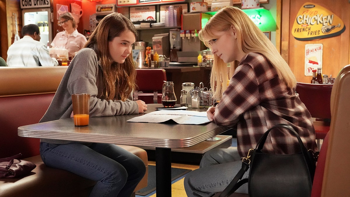 Missy (Raegan Revord) in a grey sweatshirt leans over the table as Paige (McKenna Grace) wears a plaid shirt and stares at her friend in an episode of "Young Sheldon"