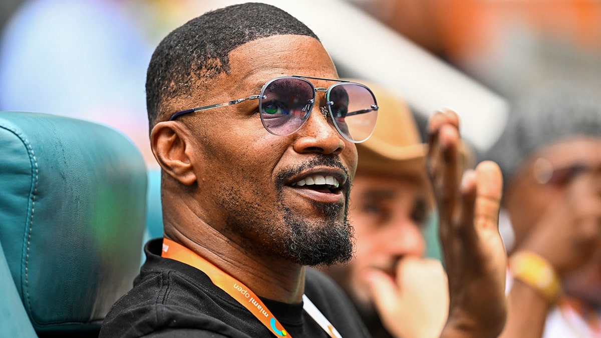 Jamie Foxx raises his hand and waves with an orange lanyard around his neck wearing a black shirt while watching tennis in Miami
