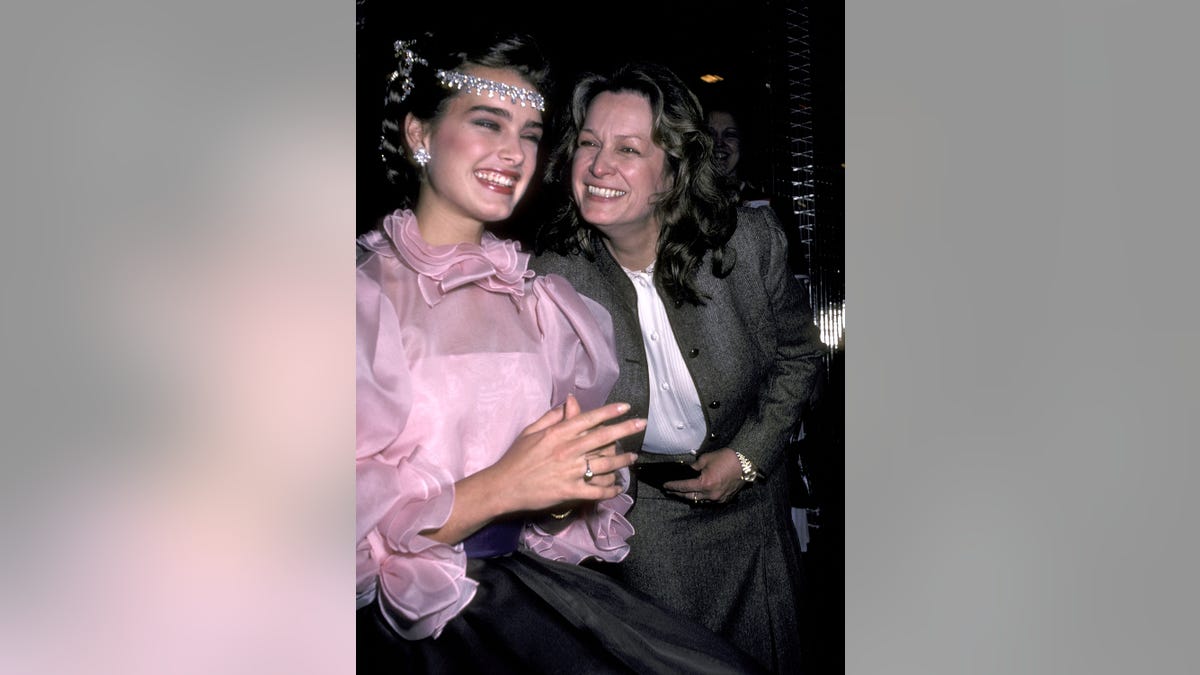 Brooke Shields smiles big in a light pink ruffled blouse and jeweled headpiece next to her mother Teri in a suit and white shirt