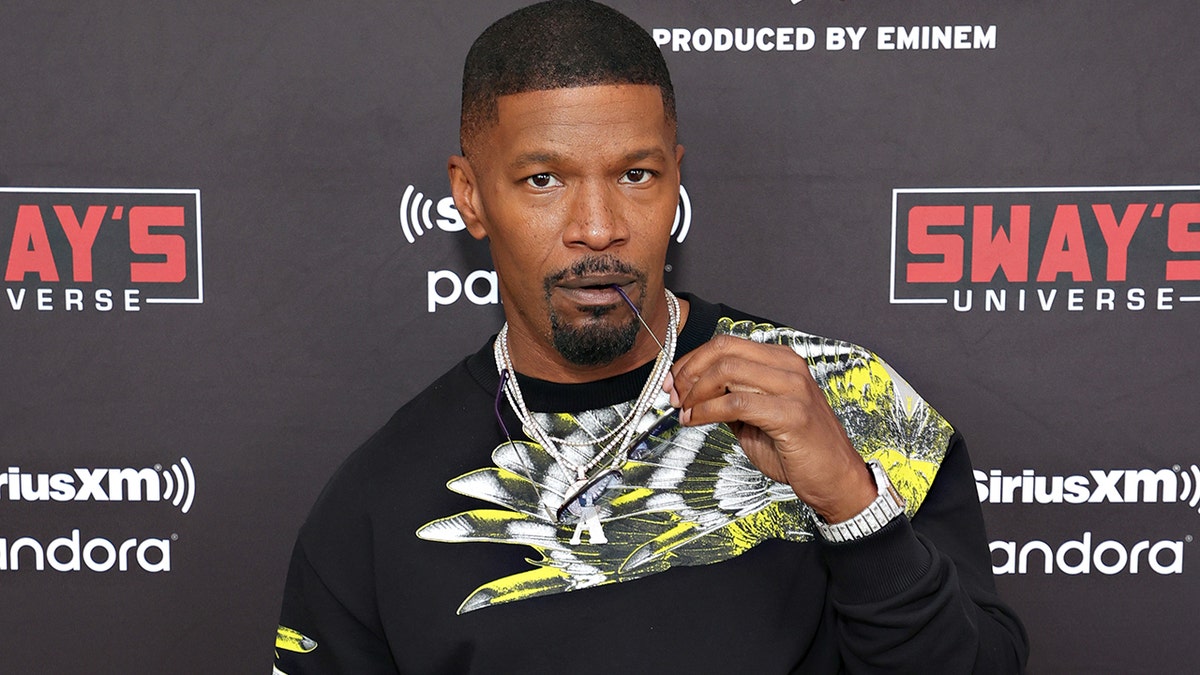 Jamie Foxx bites the end of his glasses on the red carpet with a large chain and black shirt with a white and neon yellow design