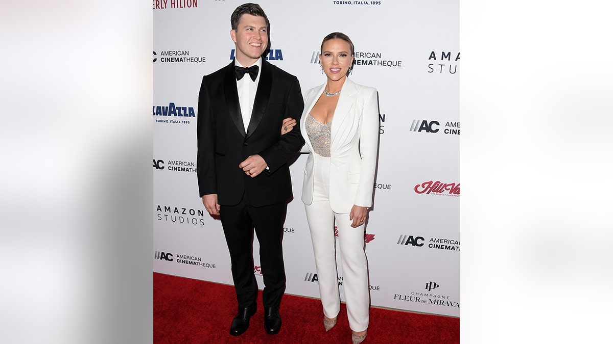 Scarlett Johansson wears a white blazer and pant set with a rhinestone bodysuit holds onto the arm of Colin Jost wearing a classic black tuxedo on the red carpet