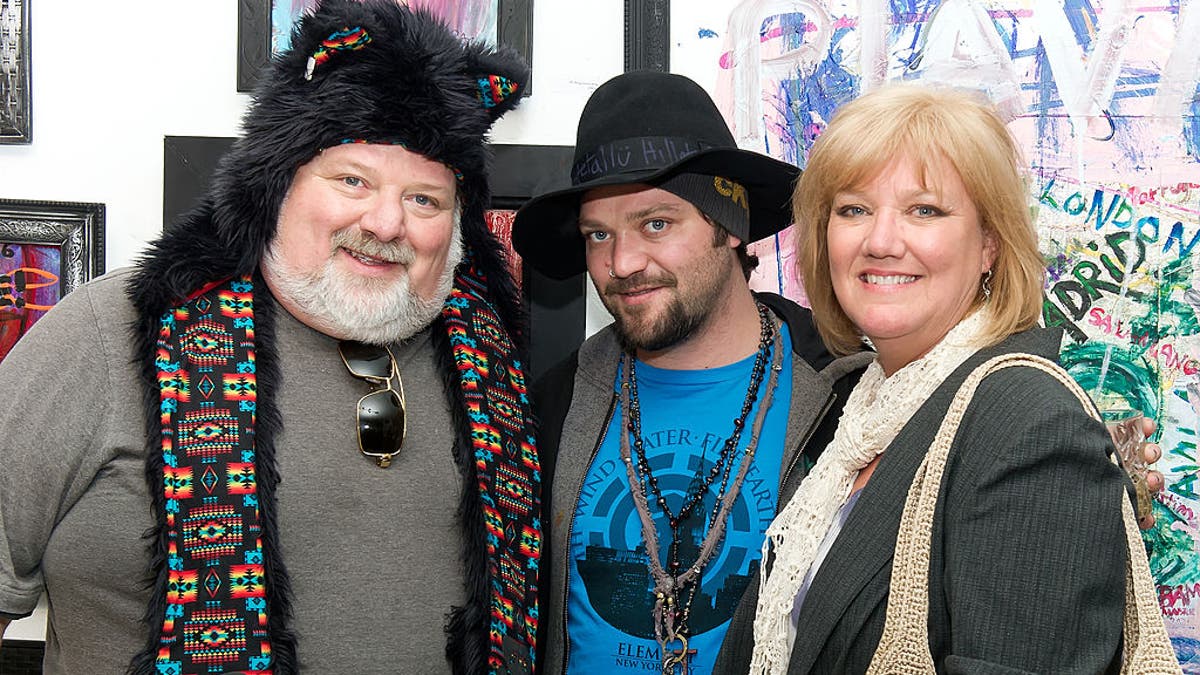 Bam Margera in a bright blue t-shirt and black brimmed hat smirks next to his father (left) Phil in a bear hat and grey shirt and mother April smiling with a scarf