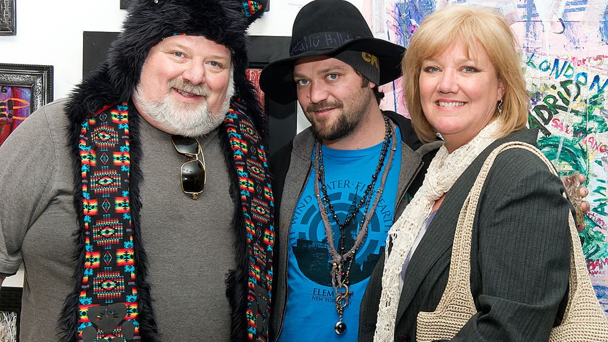 Bam Margera in a bright blue t-shirt and black brimmed hat smirks next to his father (left) Phil in a bear hat and grey shirt and mother April smiling with a scarf