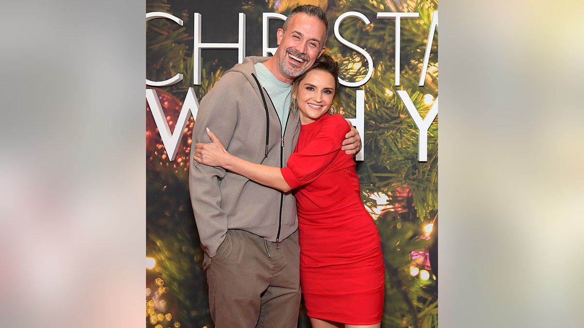 Freddie Prinze Jr. in a grey sweatshirt and light aqua blue shirt is hugged by Rachael Leigh Cook in a red dress on the red carpet for the "Christmas With You" screening