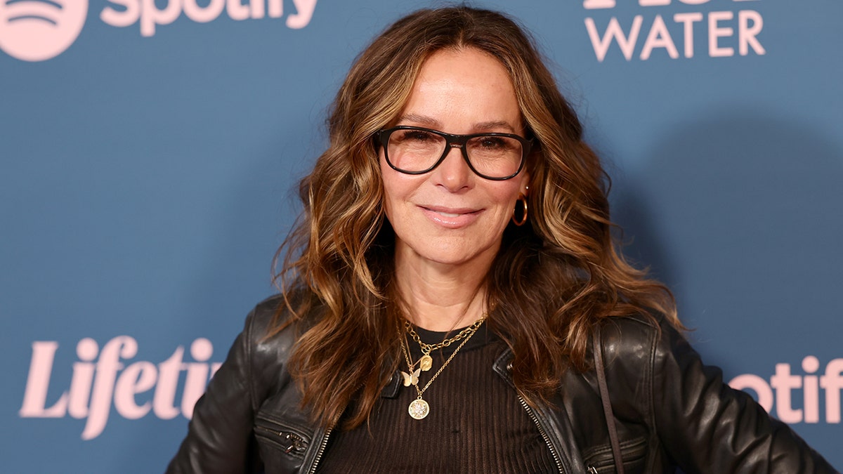 Jennifer Grey in a black leather jacket and black framed glasses smiles on the red carpet