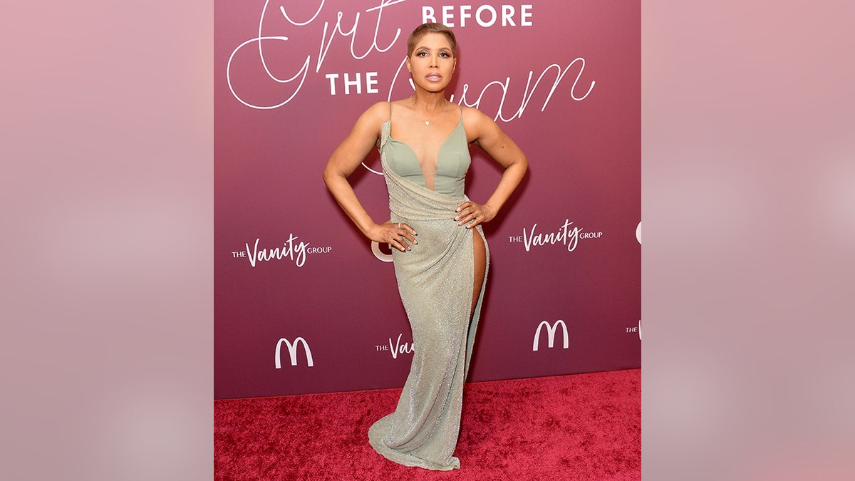 Toni Braxton has both hands on her hips in a light teal tank top with a sparkly draped skirt over it on the red carpet