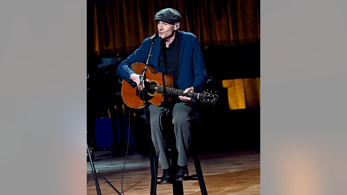 James Taylor wears a blue jacket, black shirt and pants and plays the guitar wearing a cap at the Gershwin Prize for Joni Mitchell