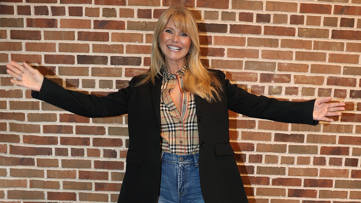 Christie Brinkley smiles with her arms stretched out wide, in a black sweater, Burberry shirt and jeans
