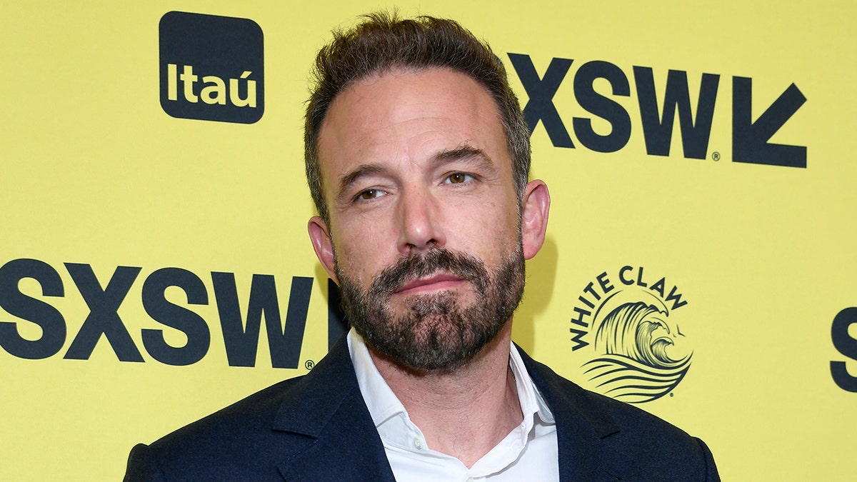 Ben Affleck looks to his right in front of a yellow SXSW backdrop, in a dark blue suit and white shirt, soft smiling