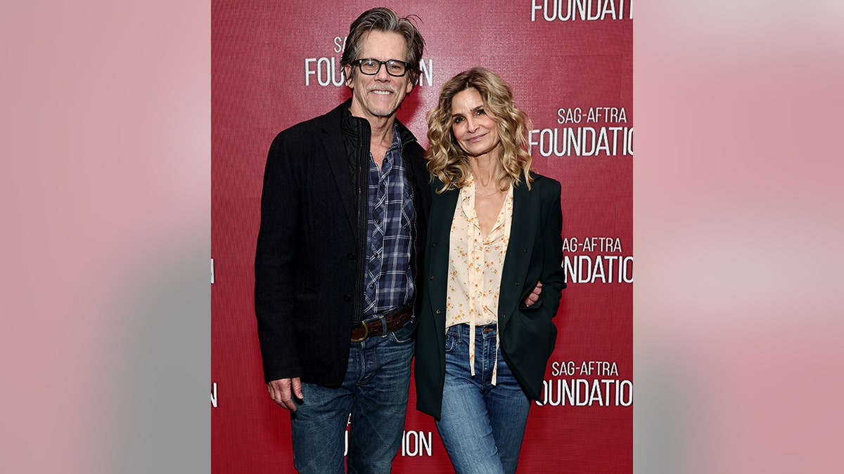 Kevin Bacon in jeans and a black jacket and blue plaid shirt smiles on the red carpet with wife Kyra Sedgwick, also in jeans, a blush blouse and black blazer