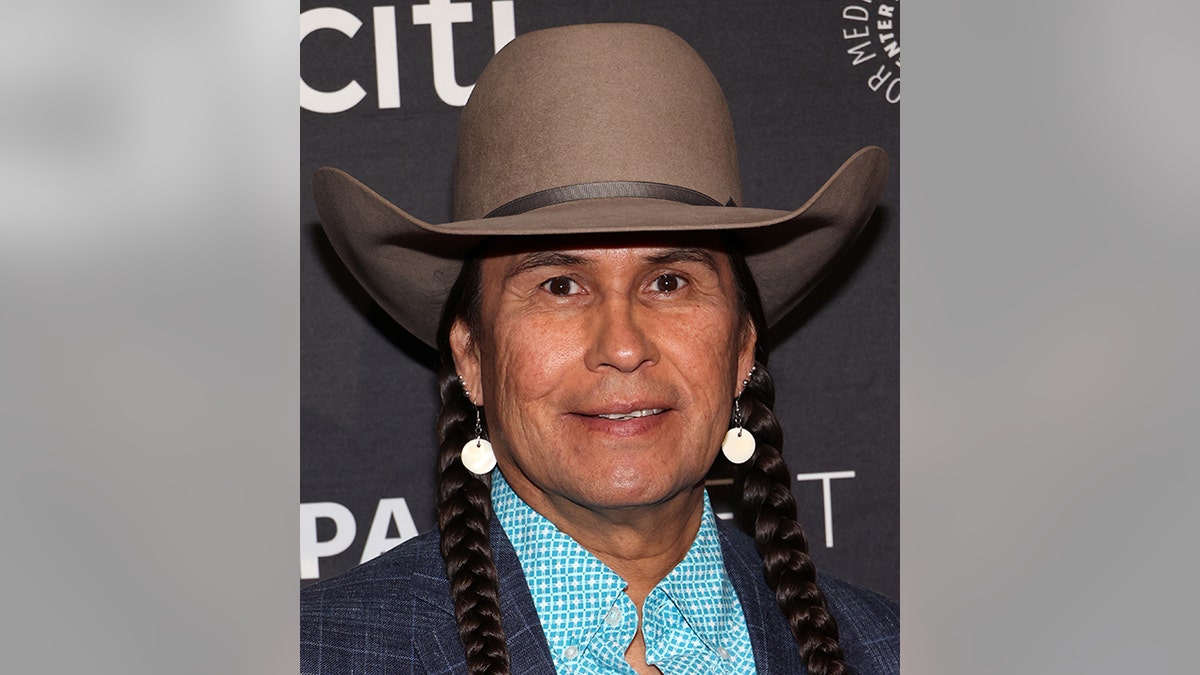 Mo Brings Plenty smiles on the PaleyFest 2023 carpet in a denim/plaid suit, turquoise patterned shirt, dangly earrings, and a large brown cowboy hat