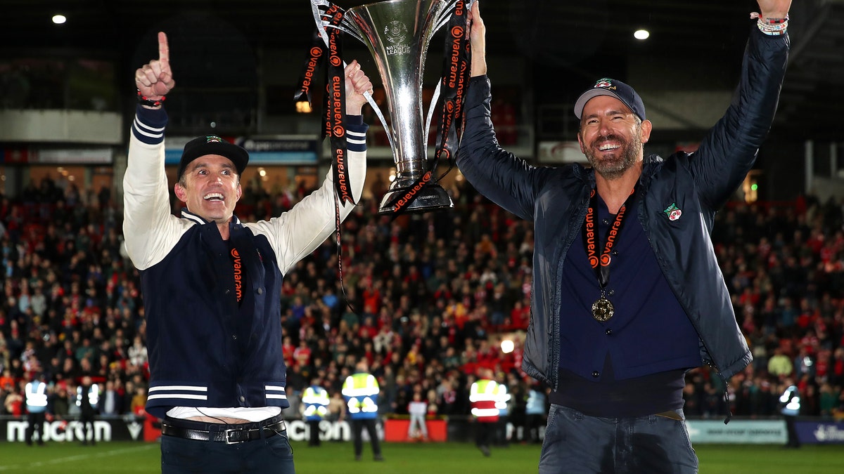 Rob McElhenney and Ryan Reynolds hold up trophy on field after Wrexham win.
