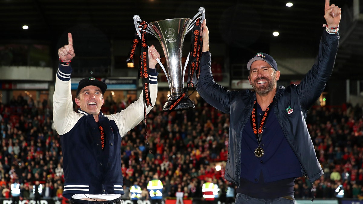 Rob McElhenney and Ryan Reynolds hold up trophy on field after Wrexham win.