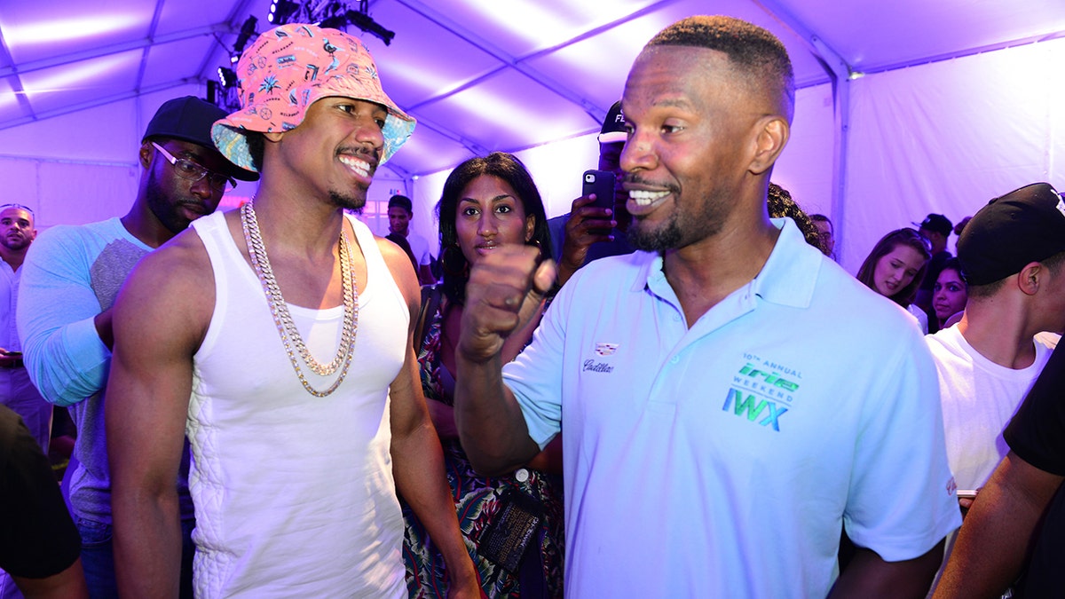 Nick Cannon in a pink hat, a white tank top and large chain smiles at Jamie Foxx who has his fist in the air wearing a blue collared shirt
