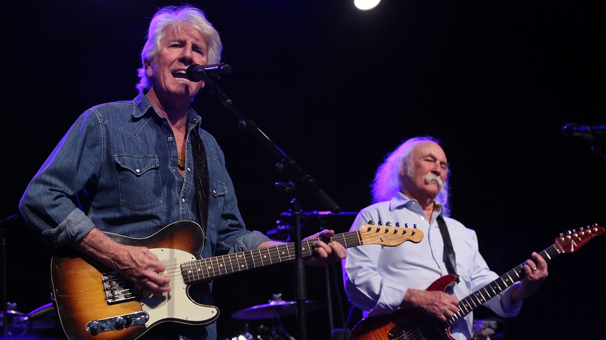 Graham Nash wears a jean shirt and sings into a microphone playing guitar in front of David Crosby wearing a light blue shirt