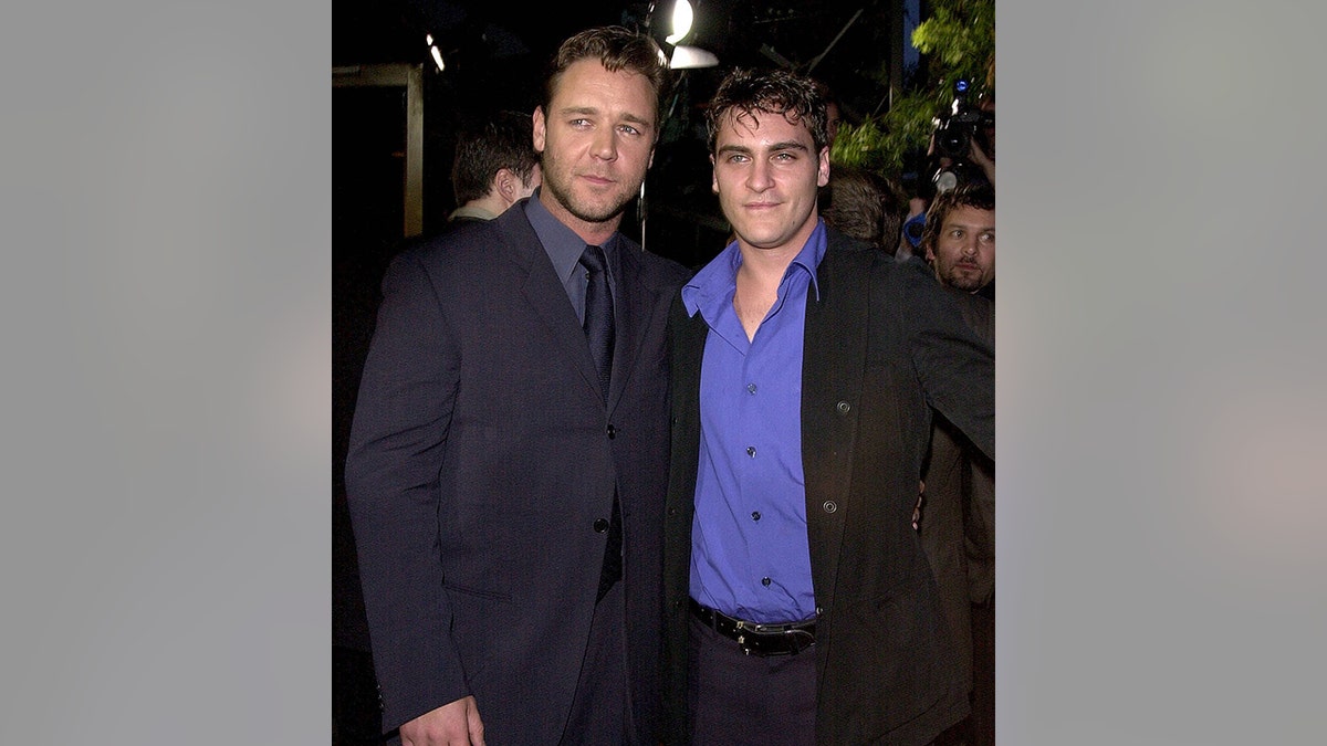 Russell Crowe in a dark suit and tie looks off in the distance to the right posing next to Joaquin Phoenix in a royal blue shirt and dark jacket