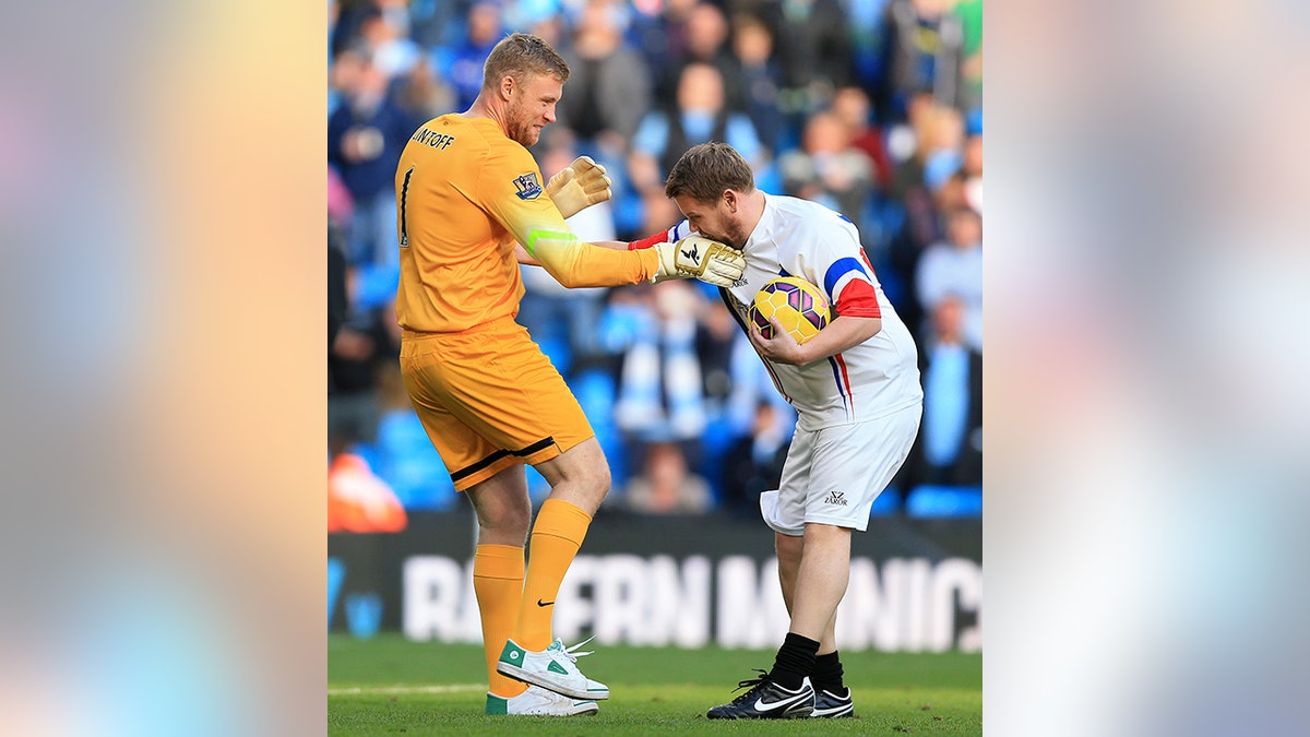 Andrew Flintoff in a dark mustard yellow shorts and shirt and cleats gets his hand kissed by James Corden in white holding a ball while filming "A League of Their Own"