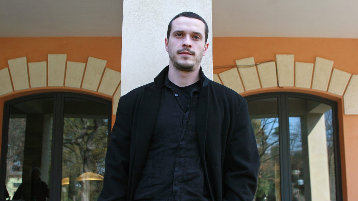 Actor Christo Jivkov wears a black coat and shirt posing in front of a building.