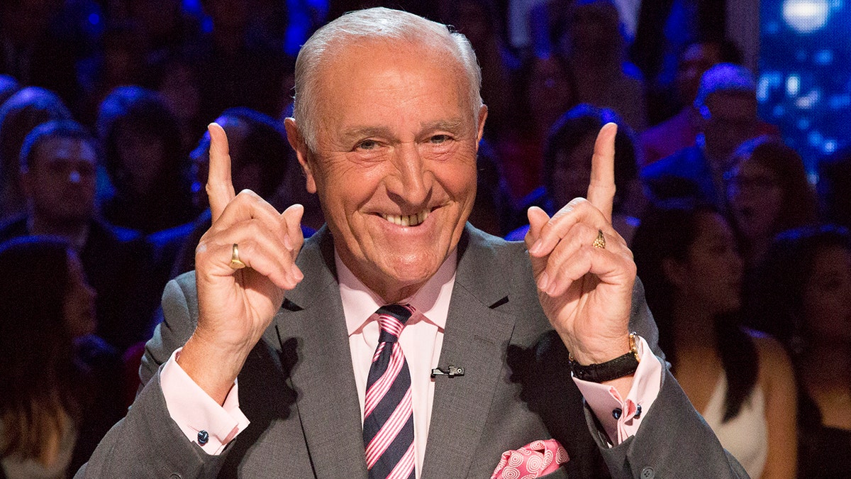 Len Goodman points both fingers up in the air, wearing a grey suit, a pink pocket square, and pink and navy blue striped tie on the set of "Dancing with the Stars"