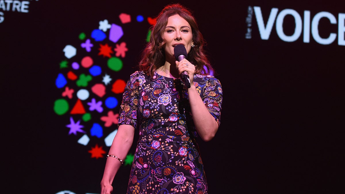 Laura Benanti sings into a microphone on stage wearing a purple floral dress