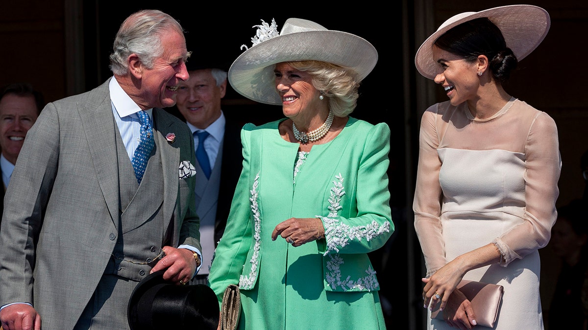 King Charles in a grey multi-piece suit smiles at Meghan Markle in a blush dress and hat, in the middle Camilla smiles in a bright green dress and white hat, looking at Charles