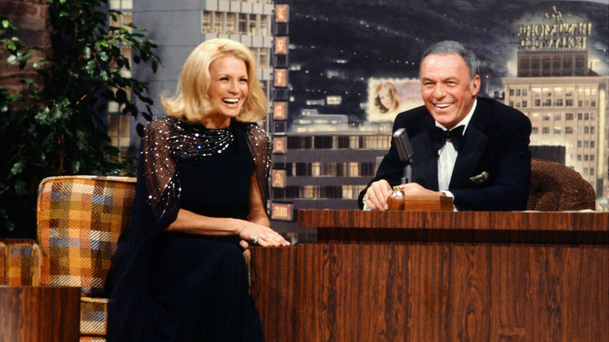 Angie Dickinson wearing a black dress and laughing next to Frank Sinatra in a suit and bow tie
