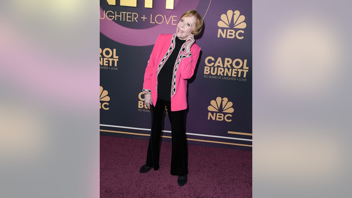 Carol Burnett wearing a black pantsuit with a hot pink blazer holding her ear