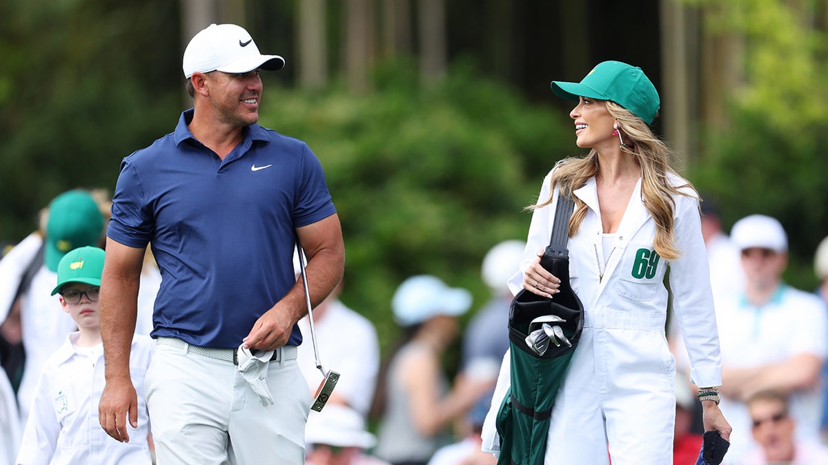 Brooks Koepka wearing white pants and a blue shirt and a white cap walking alongside his wife Jena Sims in a white caddy uniform
