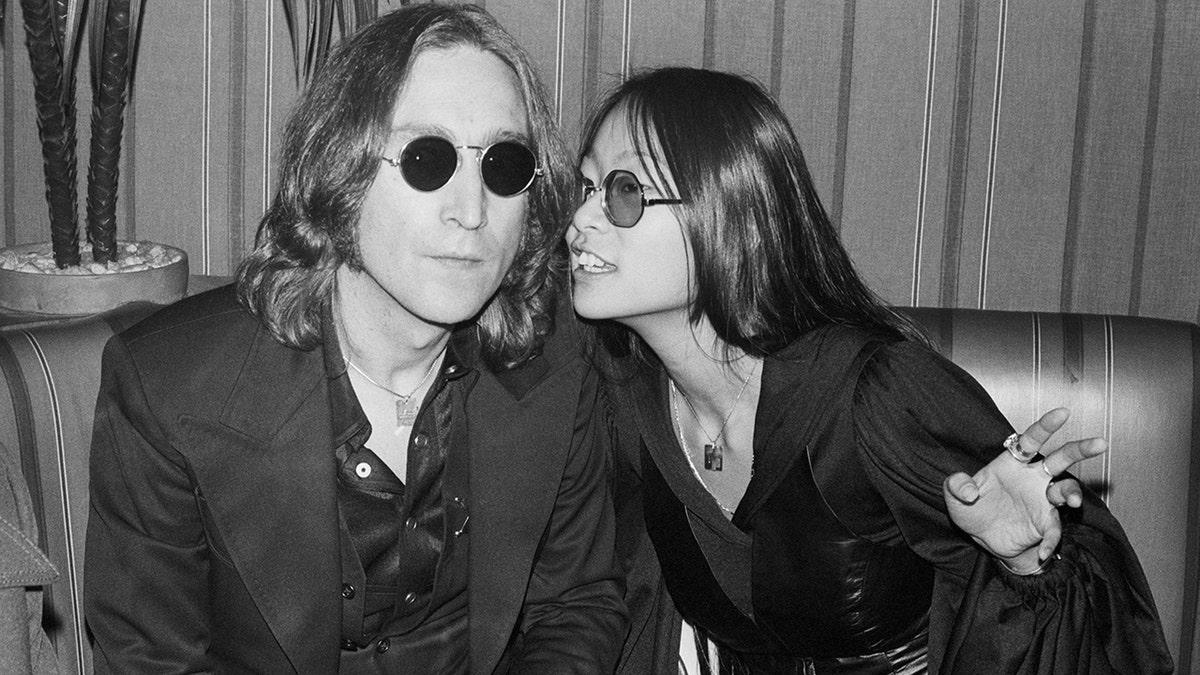 May Pang and John Lennon wearing dark clothes sitting at a restaurant