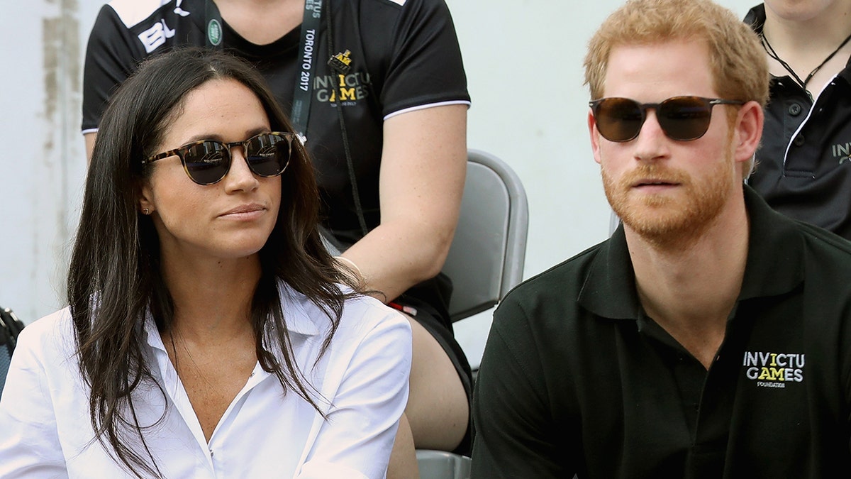 Meghan Markle wearing a white blouse with black sunglasses sitting next to Prince Harry in a black shirt and sunglasses