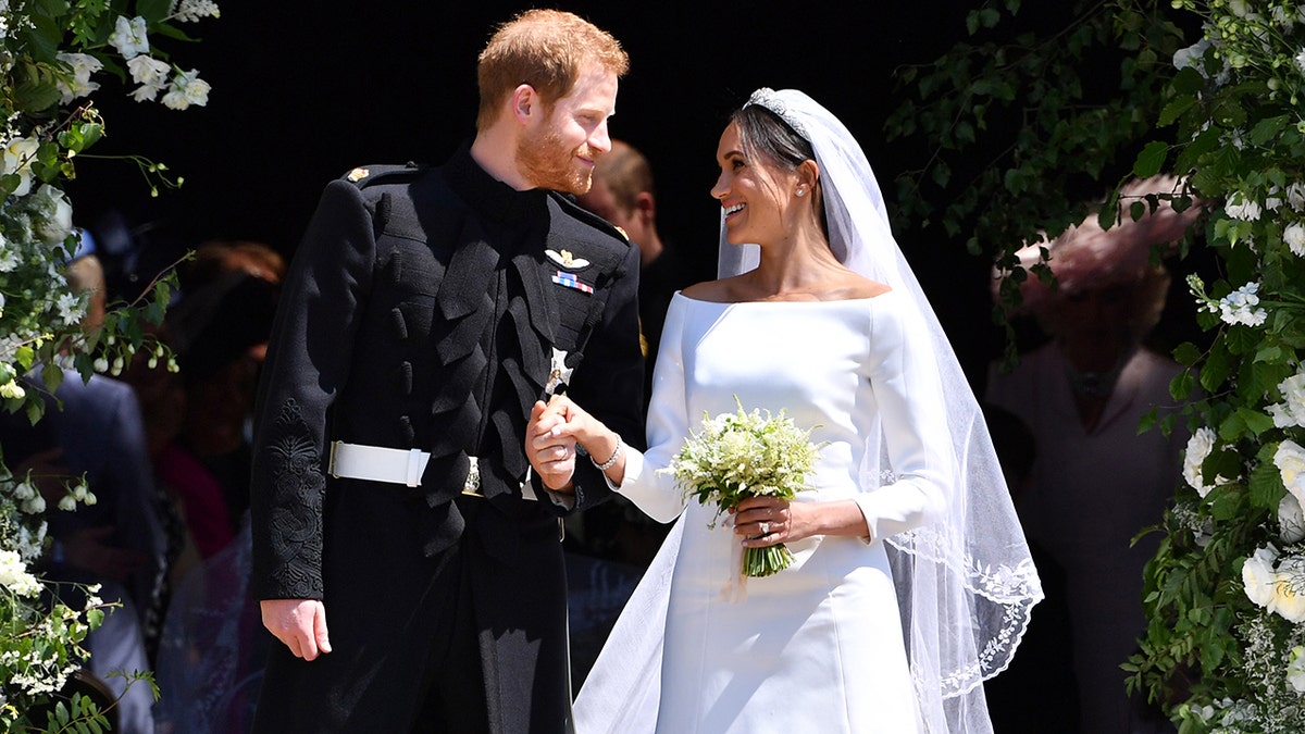 Meghan Markle wearing a white bridal dress holding Prince Harrys hand, who is wearing a suit