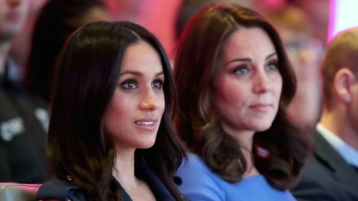 A side photo of Meghan Markle wearing a dark hued dress next to Kate Middleton wearing a blue dress