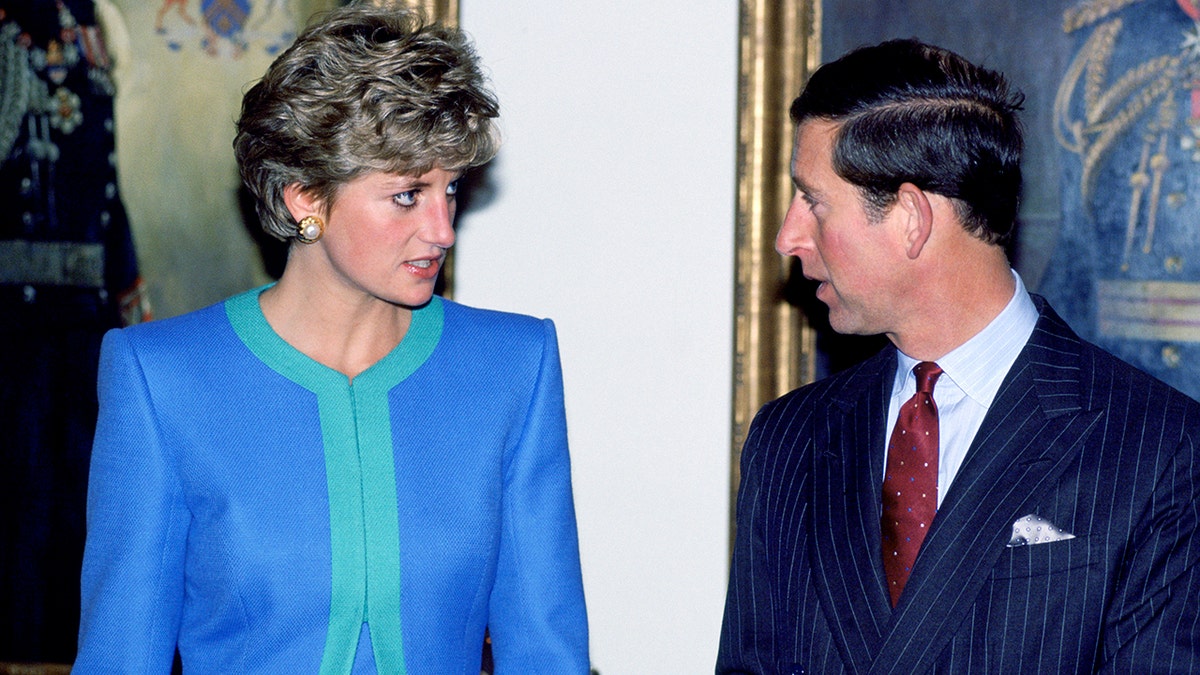 Princess Diana in a blue and green dress looking seriously at King Charles in a suit and red tie