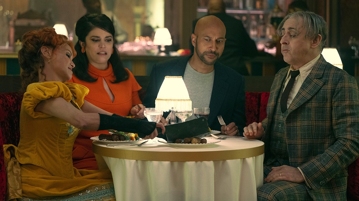 The cast of "Schmigadoon!" including Kristin Chenoweth in yellow, Cecily Strong in a bright orange, Keegan-Michael Key in a grey shirt and navy blazer and Alan Cumming in a green plaid suit set sit around a table in a photo from the second season