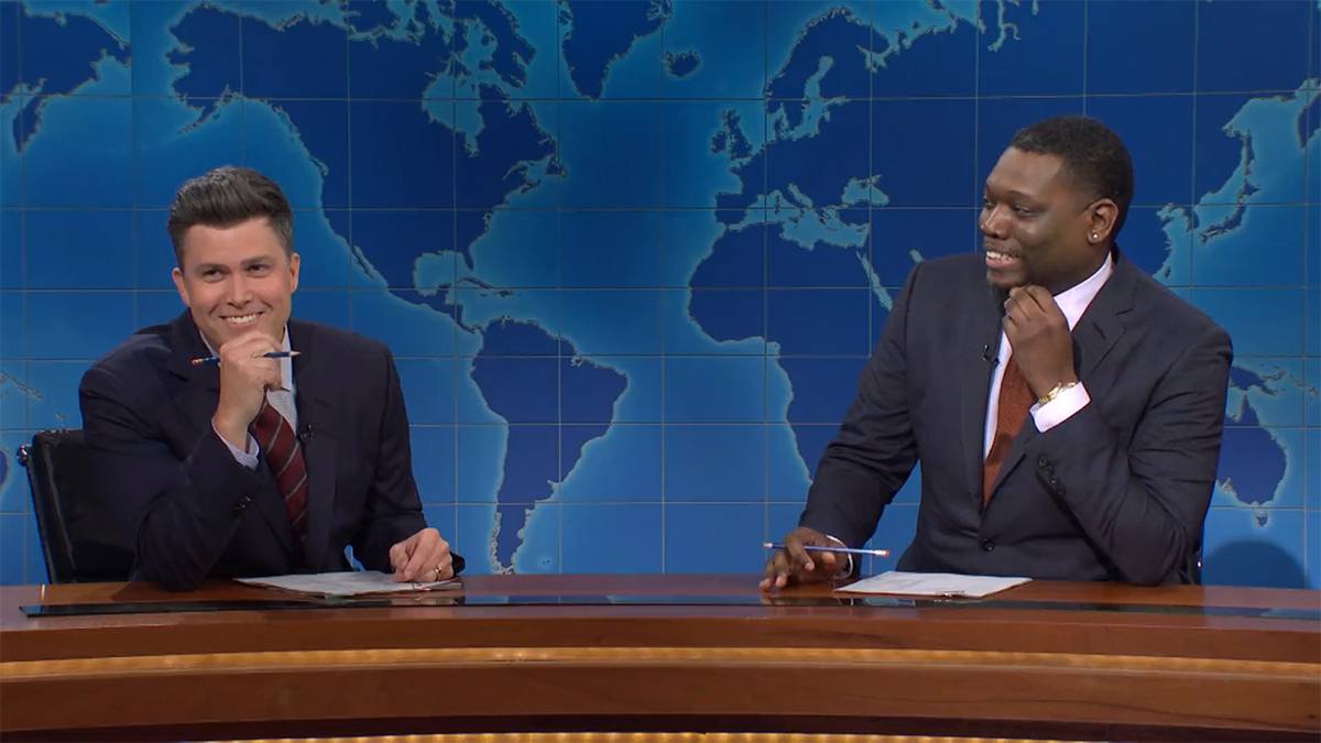Colin Jost smiles in a navy suit and dark red striped tie holding a pencil with his hand to his chin while Michael Che in a navy suit and dark orange tie looks at him with his hand to his chin