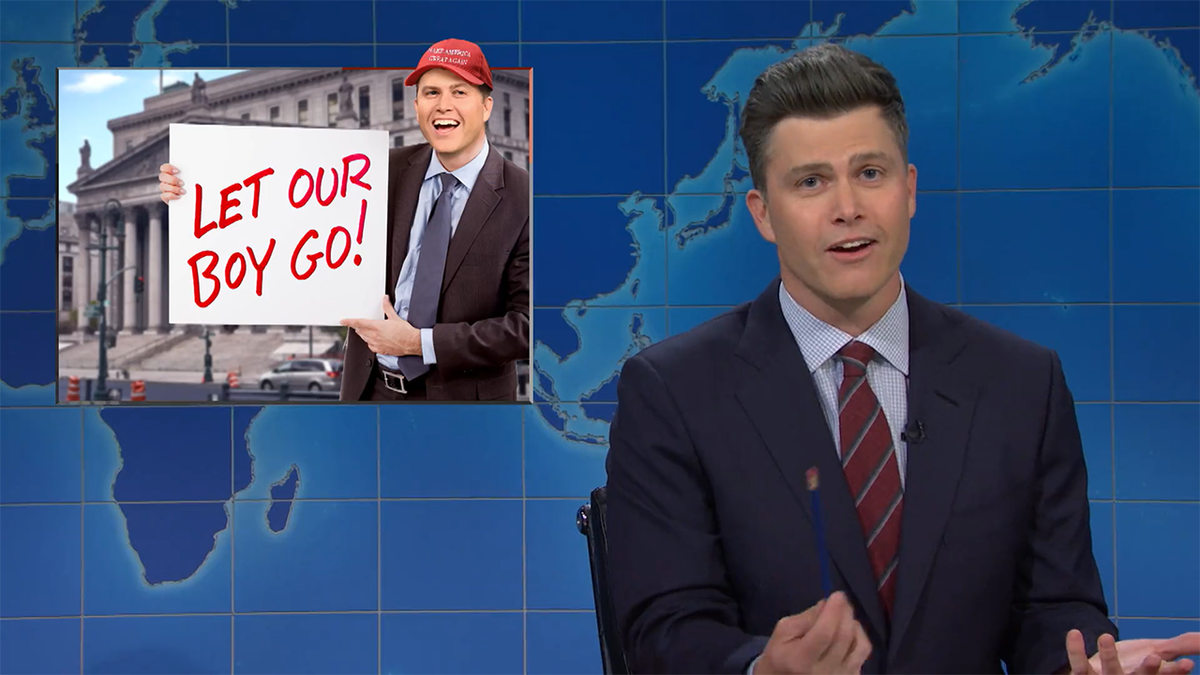 Colin Jost in a navy suit and red striped tie does his 'Weekend Update' bit on 'SNL,' inset a photoshopped image of him 'protesting' for Donald Trump