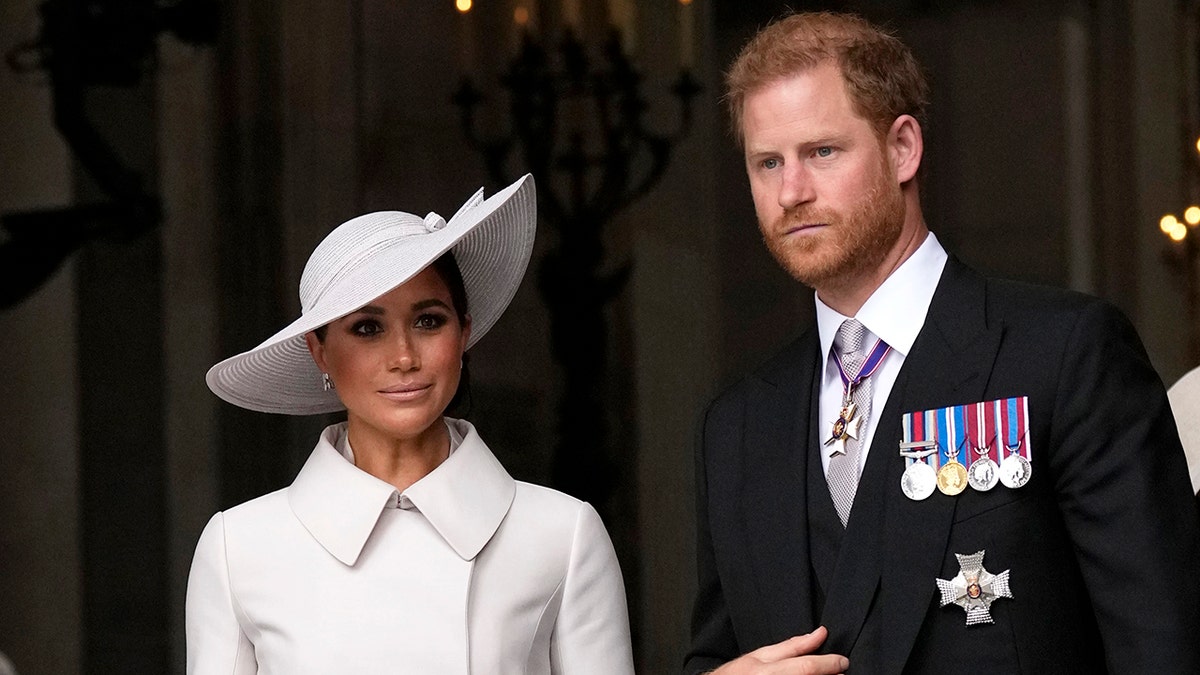 Meghan Markle wearing a white coatdress with a matching hat next to Prince Harry in a suit with his medals