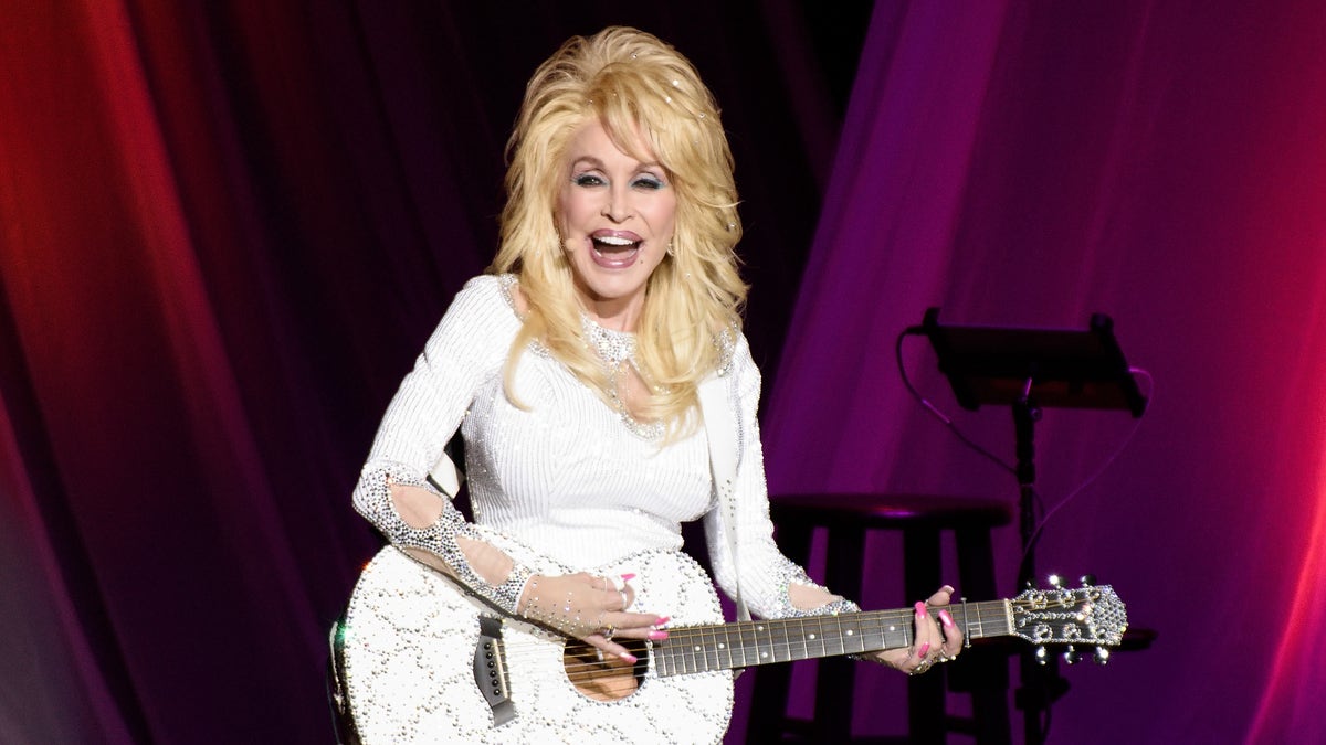 Dolly Parton wears a white ensemble and plays a white guitar.